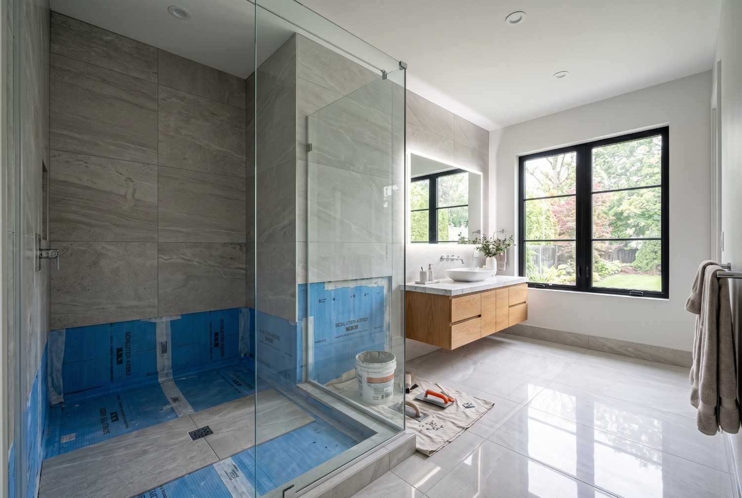 Modern bathroom renovation in progress featuring a walk-in glass shower, floating wood vanity, large-format tile, and waterproofing installation. Contemporary bathroom remodel showcasing premium finishes, custom cabinetry, natural light, and professional residential construction craftsmanship.