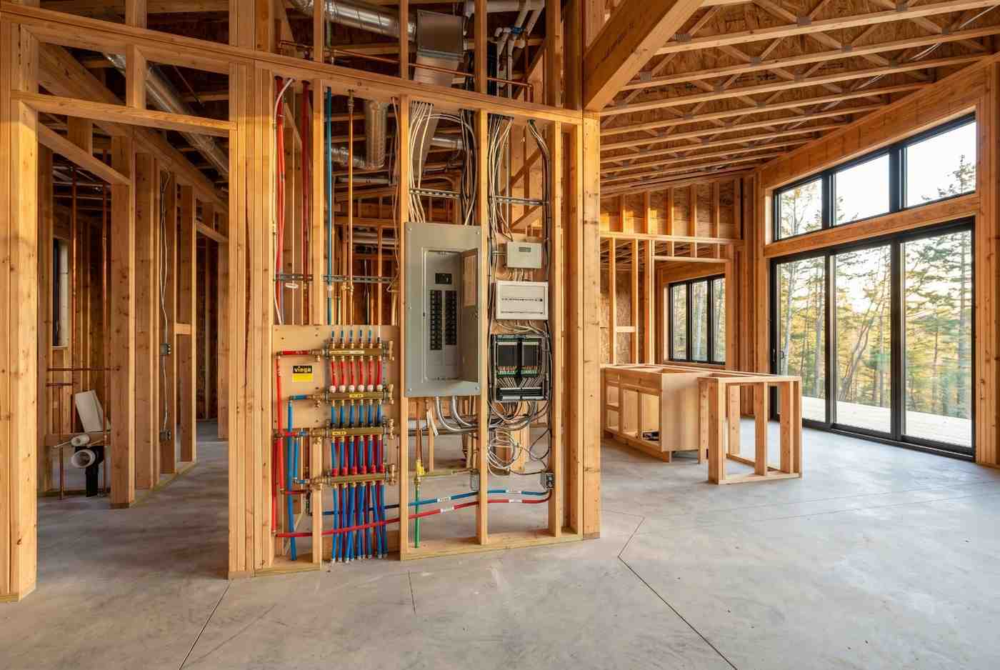 Modern home construction interior with exposed wood framing, electrical panel installation, plumbing manifold system, and open floor plan. Residential building project showcases rough-in mechanical systems, custom home construction, energy-efficient design, and large windows overlooking a natural landscape.