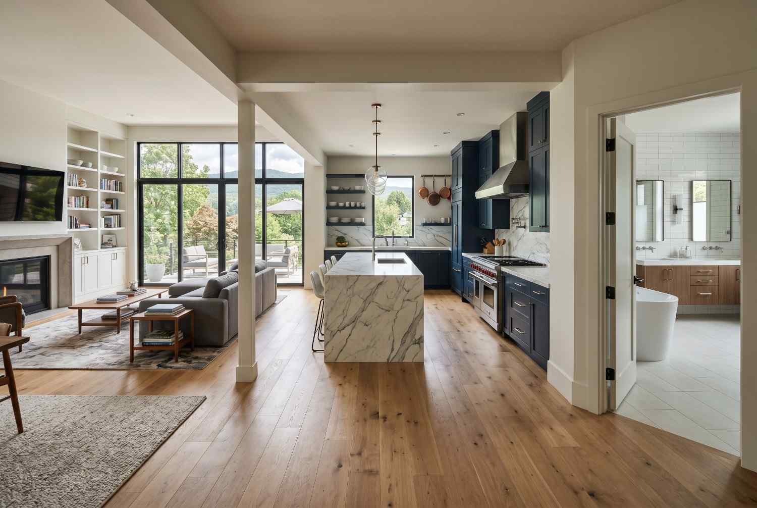 Modern open-concept kitchen, living room, and bathroom with marble waterfall island, custom navy cabinetry, hardwood flooring, and floor-to-ceiling windows. Luxury home interior featuring premium finishes, natural light, contemporary design, and high-end residential remodeling.