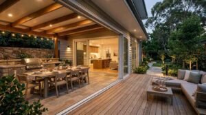 A covered patio and cedar decking area transitioning seamlessly into a modern home via open multi-panel sliding glass pocket doors. Beneath exposed timber ceiling beams strung with warm cafe lights sits a stone outdoor kitchen with a built-in stainless steel grill, sink, and prep counters, alongside an expansive wooden dining table set for dinner. The adjoining open-air deck features a contemporary cushioned L-shaped sectional sofa and coffee table with drinks, overlooking an illuminated stone pathway through landscaped evening gardens.