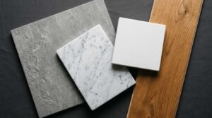 Flat lay material swatch palette featuring textured grey stone tile, white Carrara marble, solid white quartz, and natural oak wood flooring on a dark surface