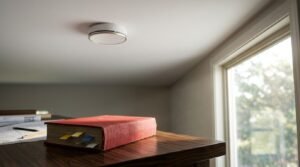 Modern round white smoke detector mounted on a white ceiling above a wooden desk with a red book, blueprints, and a window letting in natural light inside a tiny home.