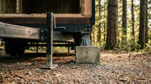 Close-up view of a tiny house trailer foundation showing an adjustable steel stabilizing jack and steel pier mounted on a concrete block foot in a wooded forest