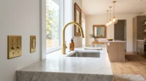 A brushed brass gooseneck faucet and undermount stainless steel sink set into a white marble kitchen countertop, beside matching brass toggle switches and a sunny open-concept interior