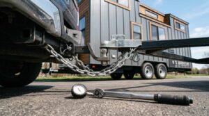 Low-angle close-up of a pickup truck tow receiver hitched to a tiny house trailer coupler with crossed galvanized safety chains, alongside a heavy-duty mechanical torque wrench and analog tire pressure gauge resting on the asphalt road