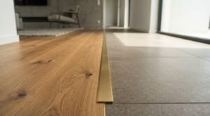 Low-angle view of a flush floor transition between natural light oak hardwood flooring and large grey porcelain tile separated by a brushed brass metal threshold strip