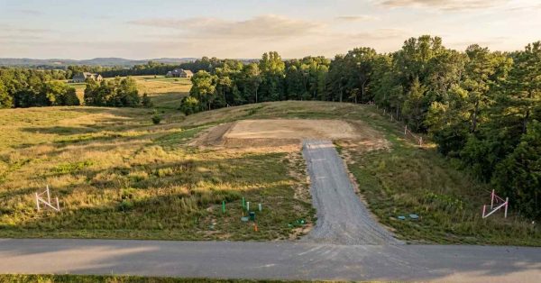 Aerial view of a vacant residential building lot with a newly graded homesite, gravel driveway access, visible utility markers, and survey boundary stakes, surrounded by open fields and mature trees.