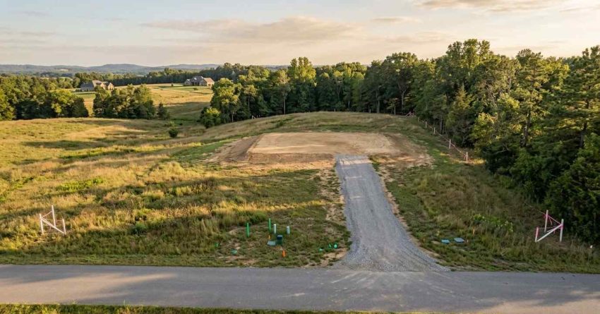 Aerial view of a vacant residential building lot with a newly graded homesite, gravel driveway access, visible utility markers, and survey boundary stakes, surrounded by open fields and mature trees.