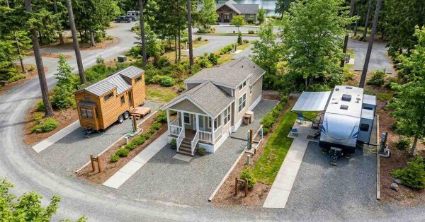 Aerial view of a planned resort community featuring a tiny home on wheels, a park model home, and a travel trailer on individual landscaped lots with utility hookups, paved walkways, and shared roads surrounded by mature trees, illustrating different compact housing options within a recreational residential setting.