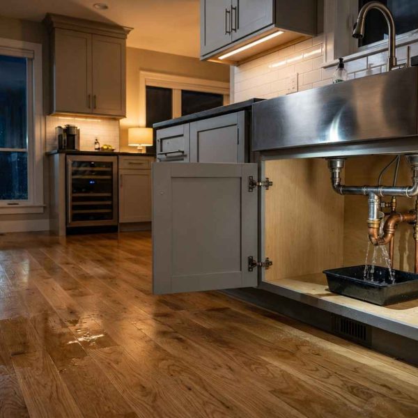 Emergency plumber repairing a leaking kitchen sink pipe in a modern home, using professional tools to stop water damage. Licensed plumbing technician fixing under-sink plumbing during a residential emergency repair service, with water on hardwood floors and homeowner nearby.
