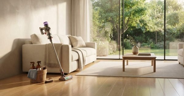 A cordless stick vacuum leaning against a neutral fabric sofa next to a wooden bucket of spray cleaning bottles on hardwood floors in a sunlit modern living room